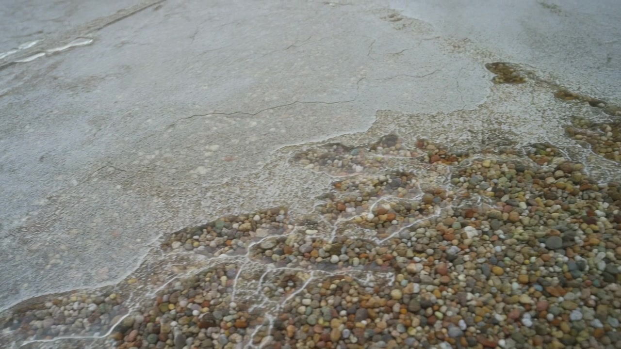 Primo piano di movimento costiero che mostra il ritirarsi dell'acqua di mare, rivelando densi agglomerati di ciottoli, pietre arrotondate sparse, sottili tracce di schiuma bianca, residui di bolle, riflessi bagnati e toni spenti e nuvolosi.