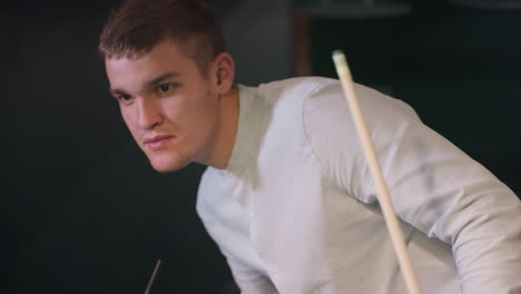 close-up of young man in white shirt with calm expression, holding cue stick and glass of lemon drink while bending to examine pool table. focused gaze and relaxed posture create a stylish