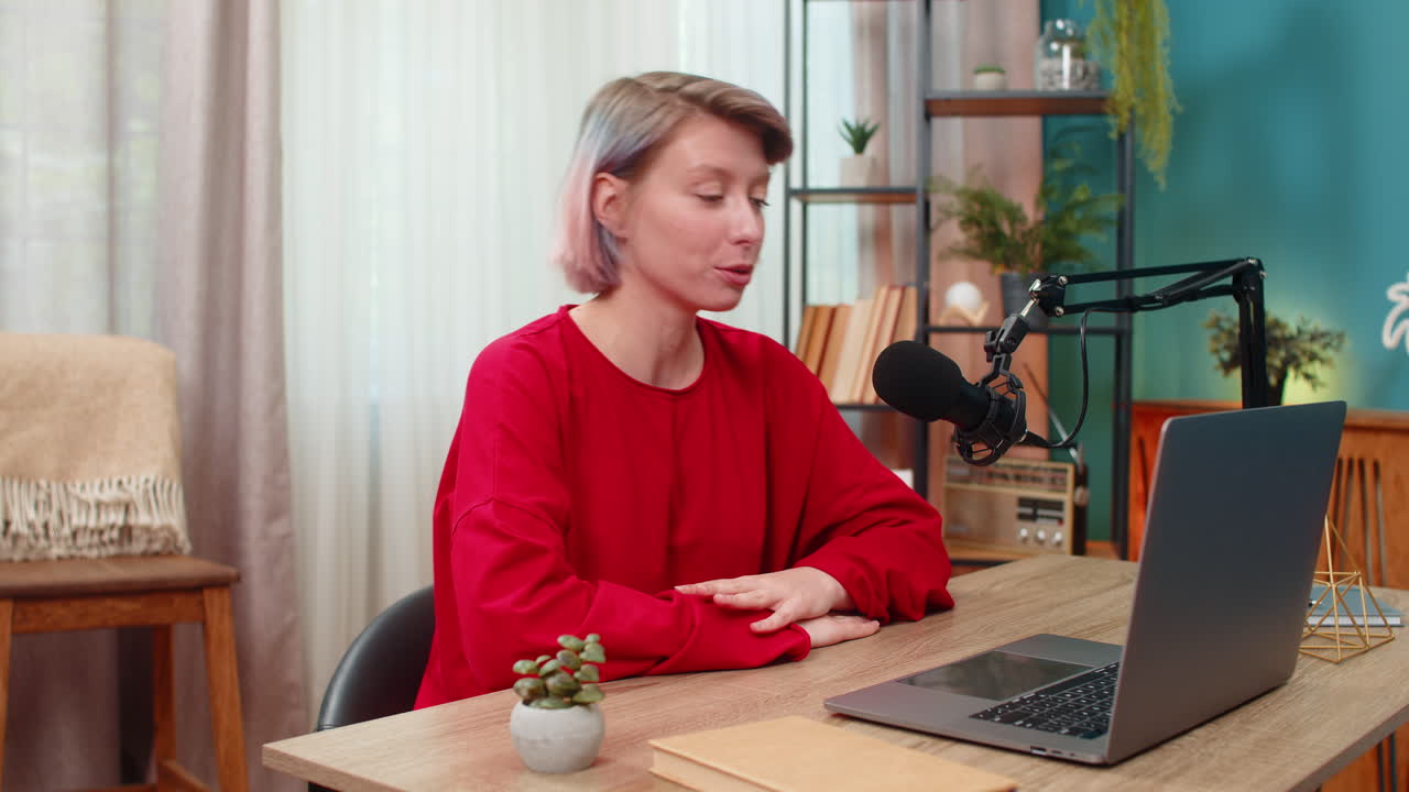 Young confident woman recording video interview at laptop speaking seriously in studio microphone