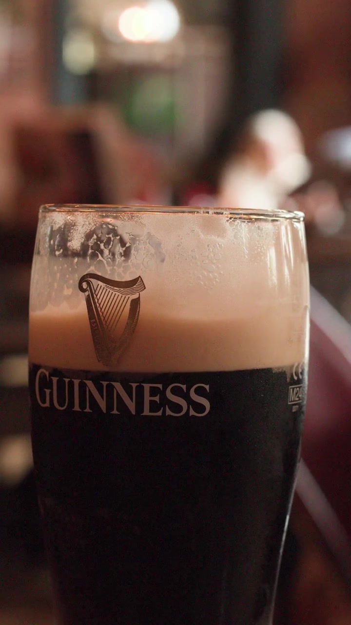 Odakta koyu renkli stout bardağı, bulanık pub iç mekanı arka planı, sıcak ortam aydınlatması, hafif kamera hareketi