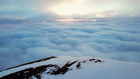 extremely long static aerial shot of a mountaineer standing on the snow covered peak of a mountain looking over an amazing clouds-cape with the setting sun on the horizon, midnight sun norway