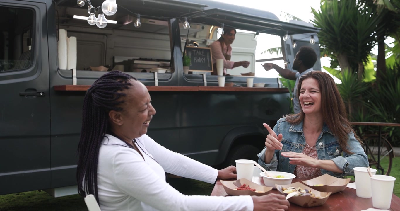 Glade multietniske kvinder, der griner og spiser ved et udendørs food truck-arrangement