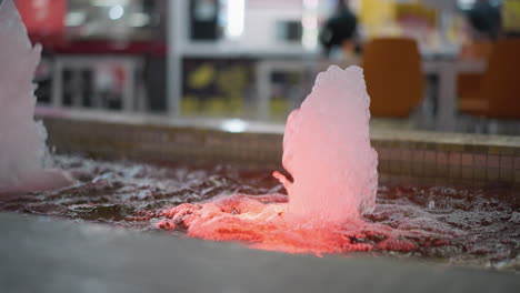 close up of water fountain with illuminated red lighting in mall, water jets creating ripples, blurred background with vibrant decor and distant shoppers