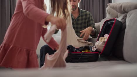 Single-father-man-brunette-wearing-a-green-checkered-shirt-together-with-his-little-daughter-brunette-girl-in-a-pink-dress-pack-their-belongings-and-clothes-in-a-black-suitcase-while-traveling-on-a-gray-sofa-in-a-modern-room