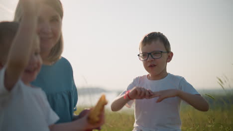 a boy in a white shirt is seen chewing and gesturing with his hands while sitting in a grassy field. in the blurred background, his mother and younger brother share a moment together