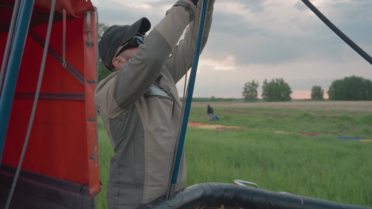 man and woman preparing hot air balloon basket, securing burner and ropes inside basket while ground crew arranges colorful envelope on green field near parked vehicles