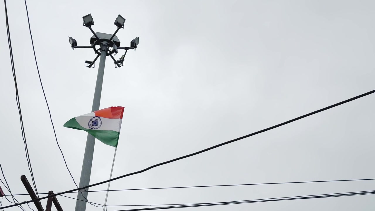 曇り空の下、頭上の電線が張り巡らされた市街地の上空に、インドの三色旗を掲げたハイマスト照明塔。