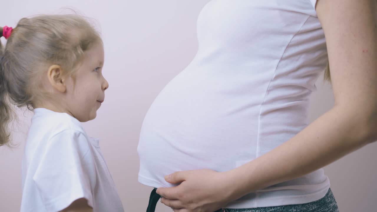 소녀는 밝은 벽에 임신 한 엄마를 안아 큰 배를 키스합니다.