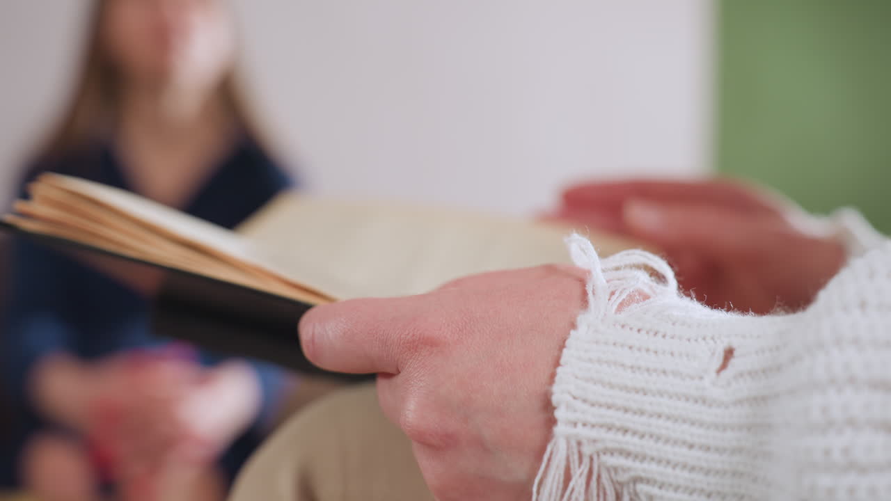Woman flipping pages of book in soft lit room while her blurred partner sits on sofa in background, evoking calm reflective atmosphere during emotional therapy session focused on healing