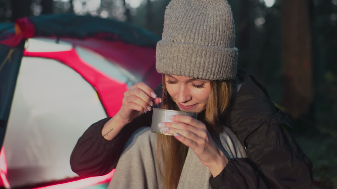 close up of lady squatting outdoors near camp tent taking bite of can food smiling relaxed casual clothing beanie forest setting evening light natural environment