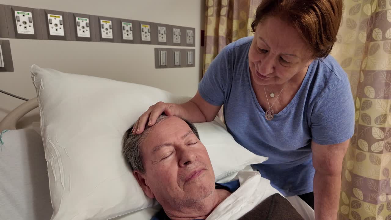 elderly man lays in the hospital bed in an unknown condition while his wife consoles him by rubbing his head