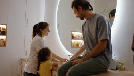 A-brunette-man-in-a-gray-T-shirt-sits-on-the-table-in-the-bathroom-and-looks-at-his-wife-and-daughter-washing-their-hands-together-in-a-modern-sink-near-the-mirror-in-a-modern-bathroom