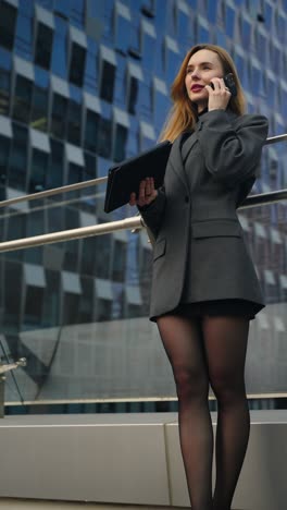 caucasian businesswoman in a gray blazer talking on phone and holding a tablet outdoors by modern office buildings - low angle parallax shot, vertical