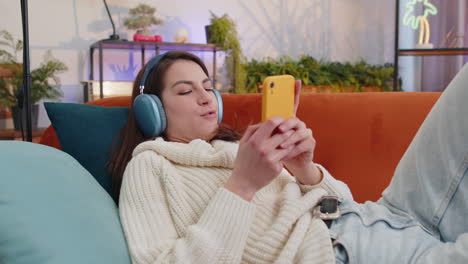 Happy-young-woman-in-wireless-headphones-relaxing-lying-on-sofa-at-home-listening-favorite-music