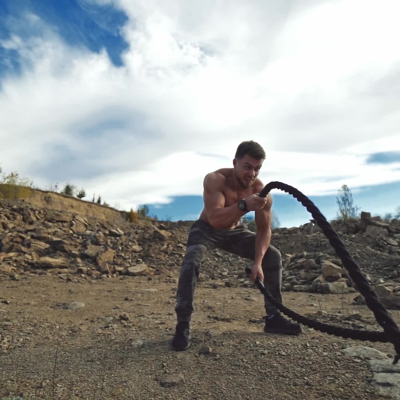 Sportsman exercising with battle ropes outdoors. Strong athlete doing double and alternating wave exercise with battle ropes standing among rocks in nature.