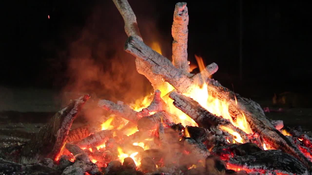 Closeup footage of burning campfire at night, glowing embers and crackling flames perfect for relaxation, camping, wilderness or cozy ambient scenes.