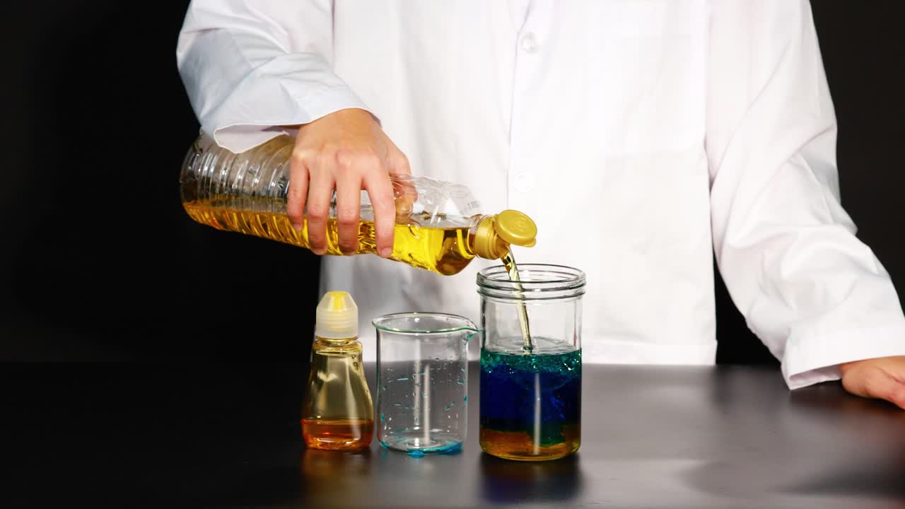 una persona con una bata de laboratorio vierte aceite en una jarra, creando capas coloridas en un experimento científico