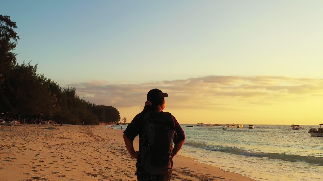 Backpacker girl walking around exotic beach at sunset with warm colors on dusty sky, alongside seashore full of fishing boats in Malaysia