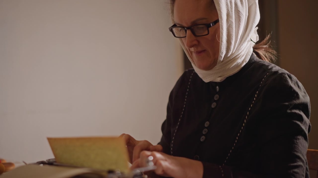 Iäkäs nainen, jolla on huivi päässään, kirjoittaa. Pehmeä, lämmin valo kylpee vintage-kirjoituskoneessa kangaspeitteisellä pöydällä. Silmälasit nenällä, keskittynyt ilme lukee kellastunutta kirjettä hitaasti ja harkitusti.
