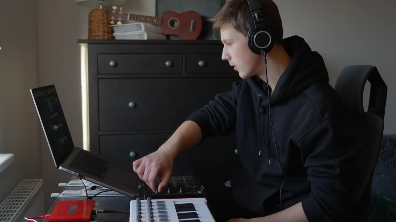 Fixed shot of a man wearing headphones, adjusting controls on a MIDI controller beside a laptop and keyboard in a home studio. A ukulele is visible in the background as he focuses on creating music
