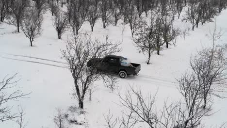 Schwarzer-Pickup-Truck,-Der-Im-Winter-Durch-Verschneite-Straße-Mit-Kahlen-Bäumen-Fährt---Antenne-Seitlich