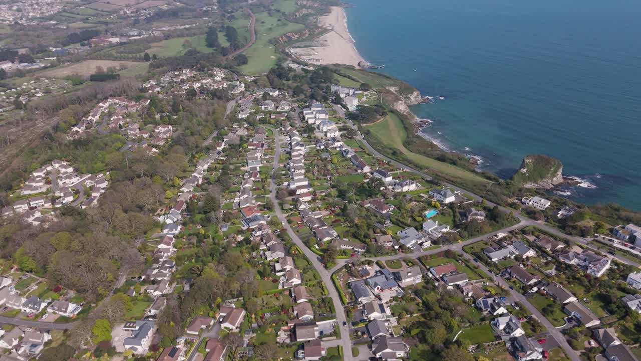 Top-down aerial of concentric residential streets where detached garden homes overlook sweeping sandy bay and wooded hills under soft coastal light