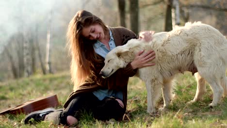 smiling happy woman gently stroking white dog around fire place.real friends people outdoor camping tent vacation in autumn trip.fall sunny day in nature, animal friendship. 4k video