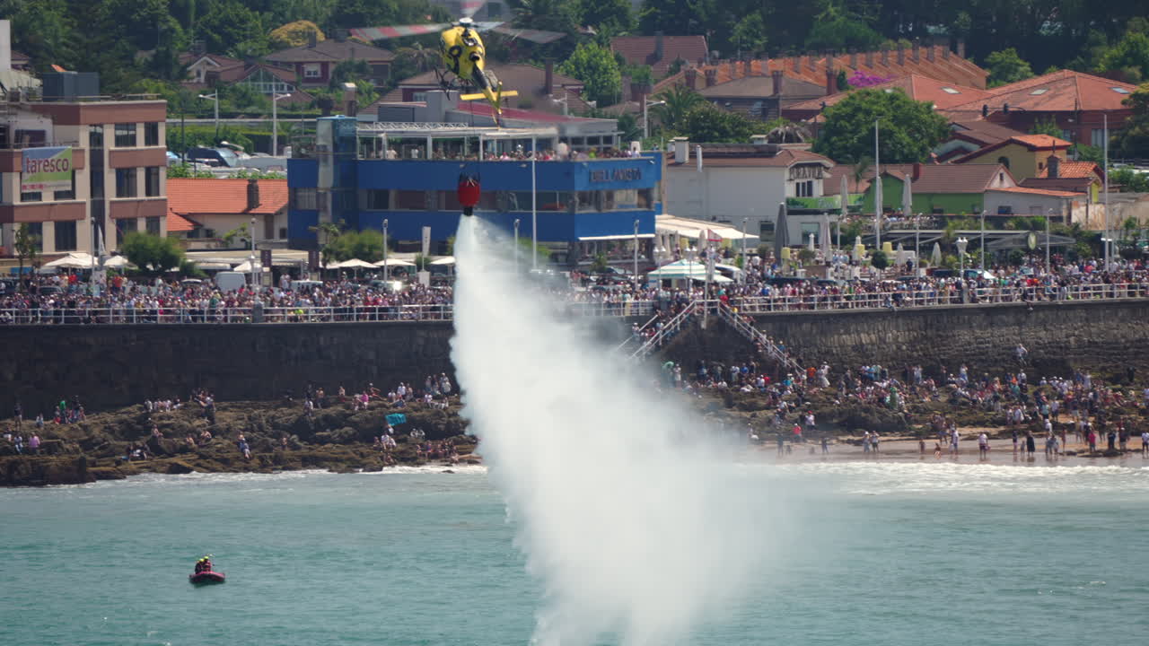 GIJON, SPAIN - JULY 27, 2025: A yellow helicopter releases a large amount of water during a firefighting demonstration at the Gijon airshow. Crowds watch from the coastline promenade as emergency aircraft perform water drops
