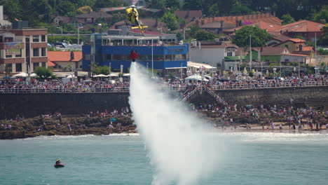 gijon, spain - july 27, 2025: a yellow helicopter releases a large amount of water during a firefighting demonstration at the gijon airshow. crowds watch from the coastline promenade as emergency aircraft perform water drops
