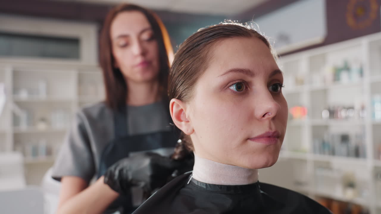 cliente seduta tranquillamente mentre lo stylist esegue il trattamento sullo sfondo, donna caucasica con mantellina e guanti protettivi, scaffalature pulite del salone e luce naturale soffusa, momento di relax durante la colorazione dei capelli