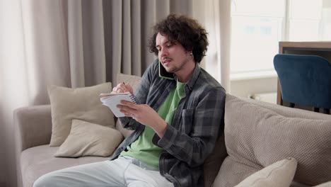 Confident-brunette-man-with-curly-hair-in-a-gray-checkered-shirt-communicates-on-the-phone-and-takes-notes-in-a-notepad-at-home-on-the-sofa