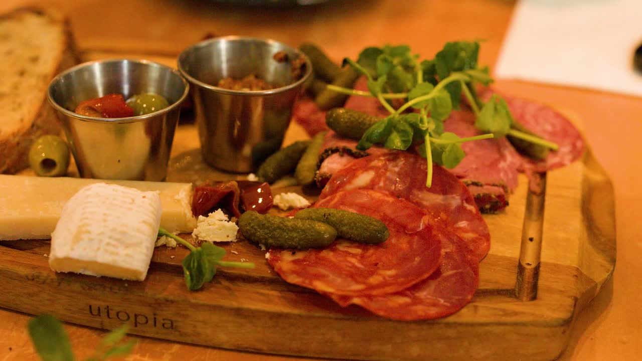 Hand slices cured meat on wooden charcuterie board with cheese, pickles, warm indoor lighting