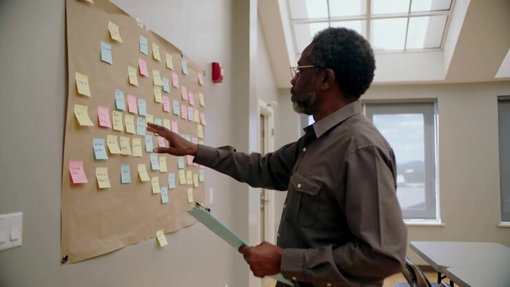 Entering meeting room, senior man holding clipboard scanning bright sticky notes on board, sorting