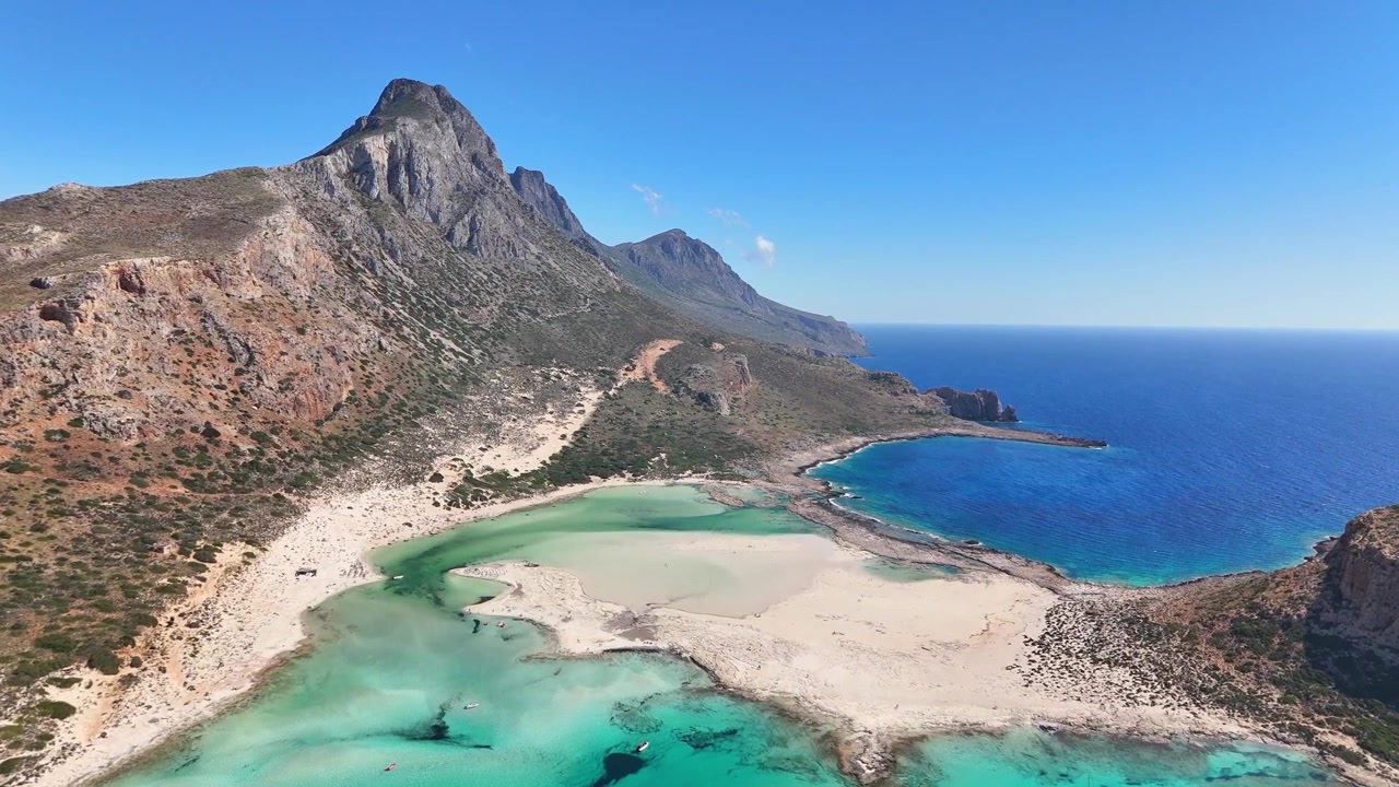 La telecamera aerea attraversa la Laguna di Balos verso una ripida montagna rocciosa, passando sopra piane di sabbia chiara, piscine turchesi poco profonde e il profondo Mar di Creta in pieno giorno.