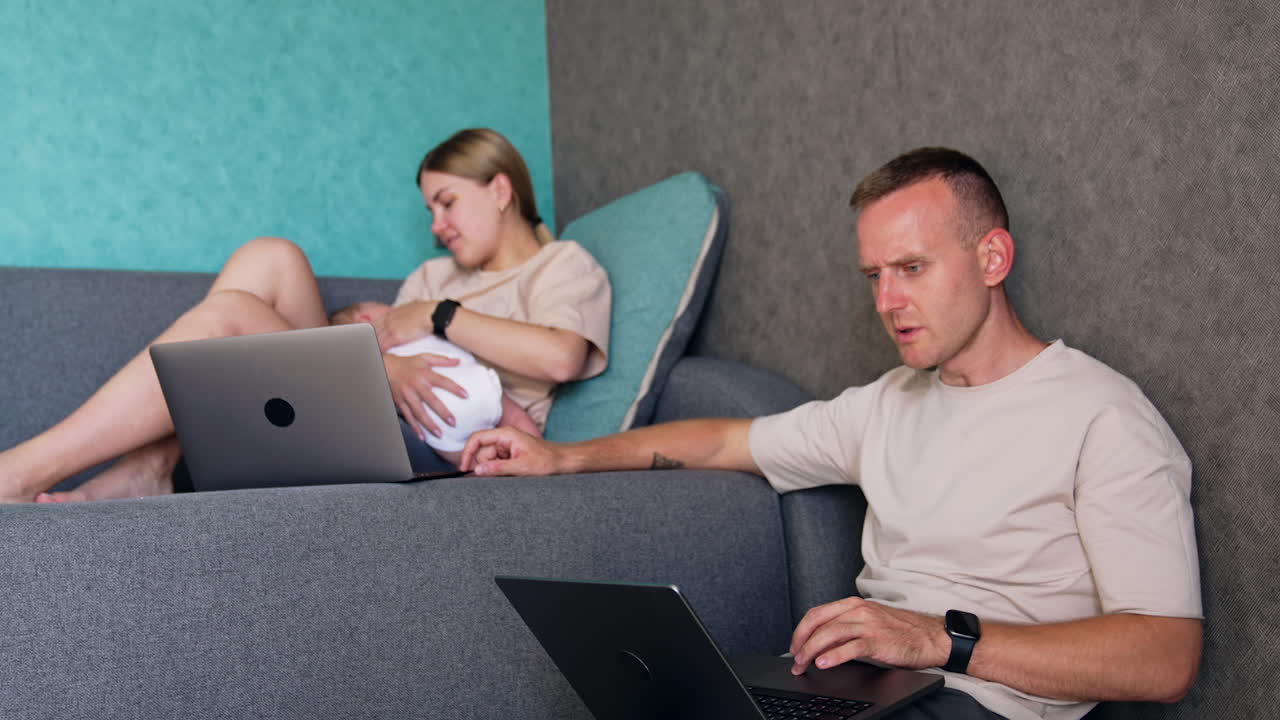 Man sitting near the sofa is focused on laptop. Woman with newborn in arms has laptop nearby and listening to a husband.