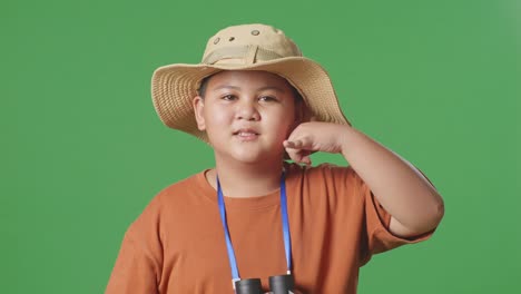 asian tourist boy with a hat and binoculars using the magnifying glass then making call me gesture on the green screen background. boy researcher examines something, travel adventure, close up