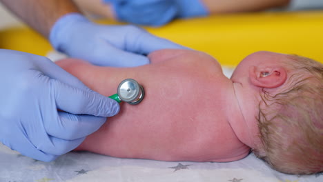 obstetrician's hands in gloves using stethoscope to examine newborn. doctor attaches medical device to a child's back. close up.