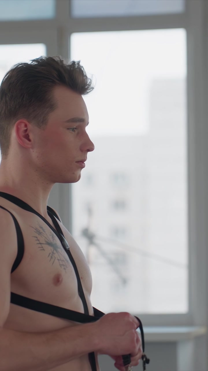 Giovane uomo a torso nudo con tatuaggi regola le bretelle nere. L'uomo tiene le cinghie vicino al collo e al petto mentre la mano di un altro uomo. La scena è ambientata da grandi finestre che lasciano entrare una luminosa luce naturale