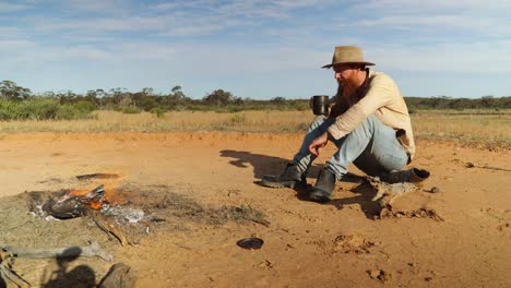 an authentic australian bushman drinks his tea from a billy by a campfire in the outback