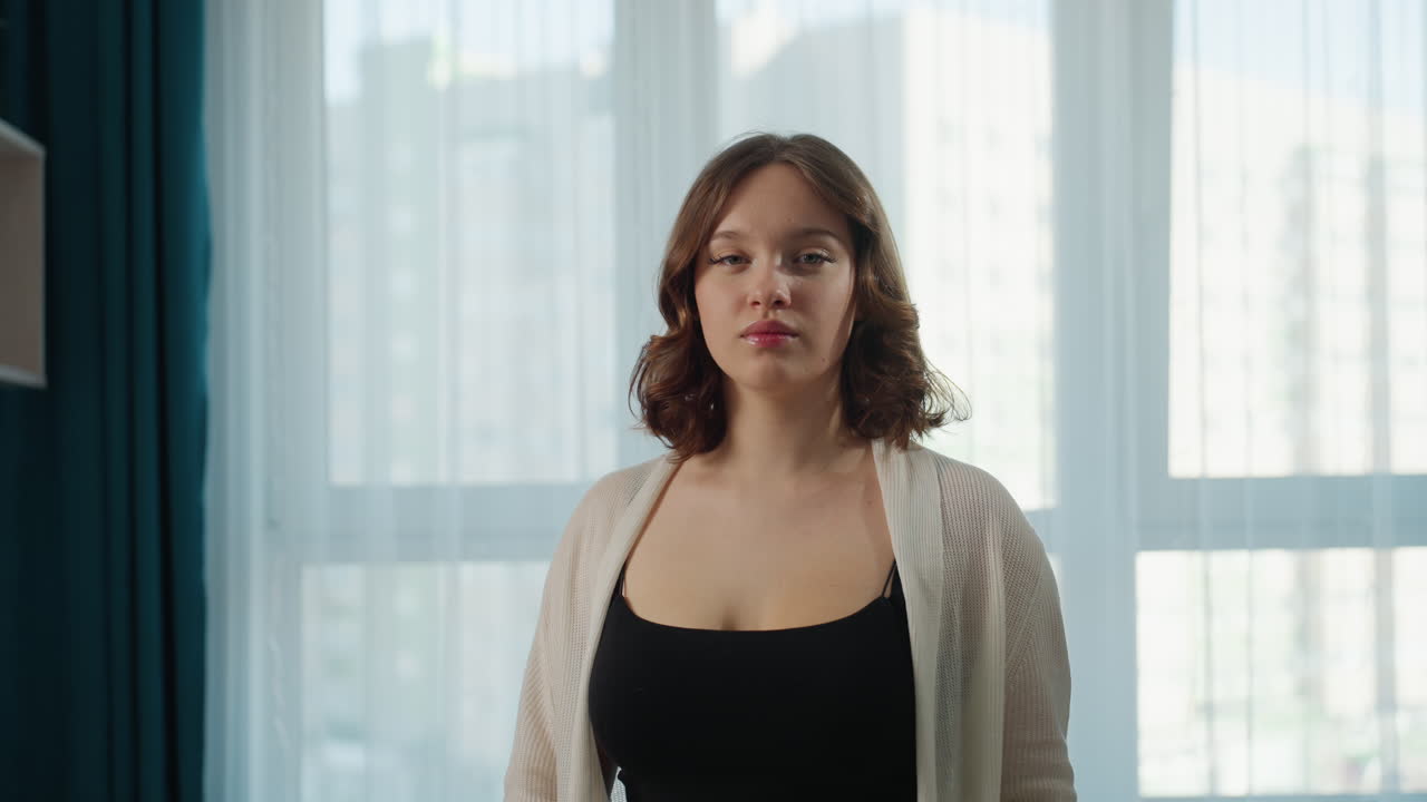 Caucasian Woman Adjusting Hair By Window, Preparing For Day, Poised Stance With Black Tank And Light Cardigan, Urban Skyline Through Sheer Curtains, Confident Expression, Selfcare Ritual, Minimal