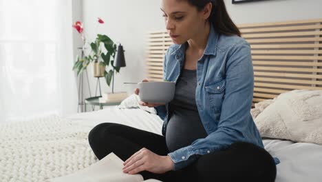 caucasian woman in advanced pregnancy sitting on bed, eating salad and reading book.