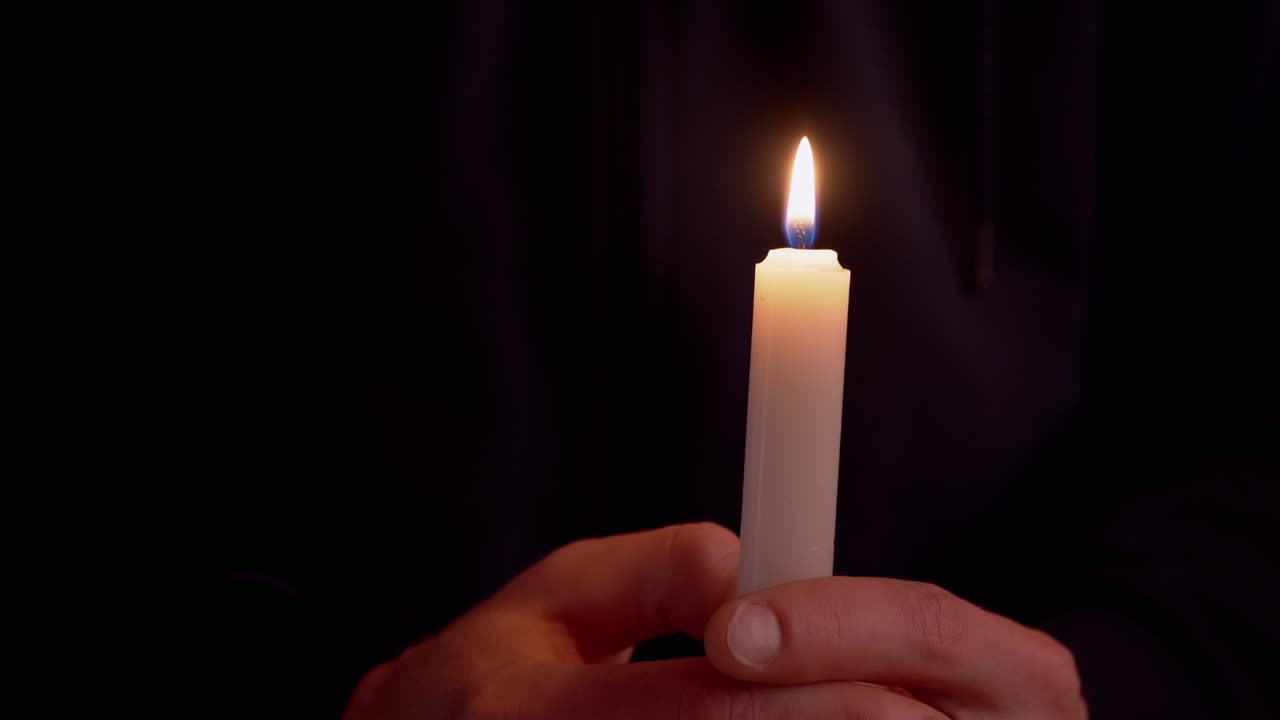 Muž drží v rukou zapálenou voskovou svíčku v tmavé místnosti. 4K.
