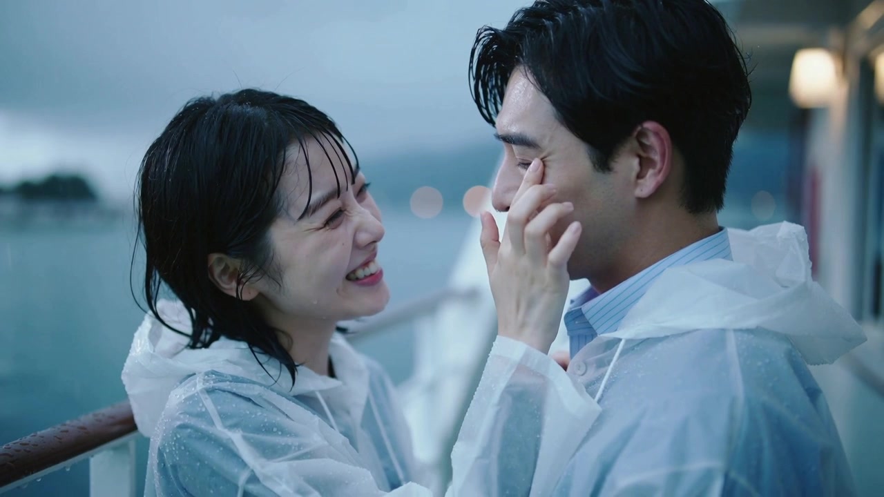 笑っている女性がシャツの襟を引っ張り、ボートのデッキで男性の頬から雨を拭き取っている。