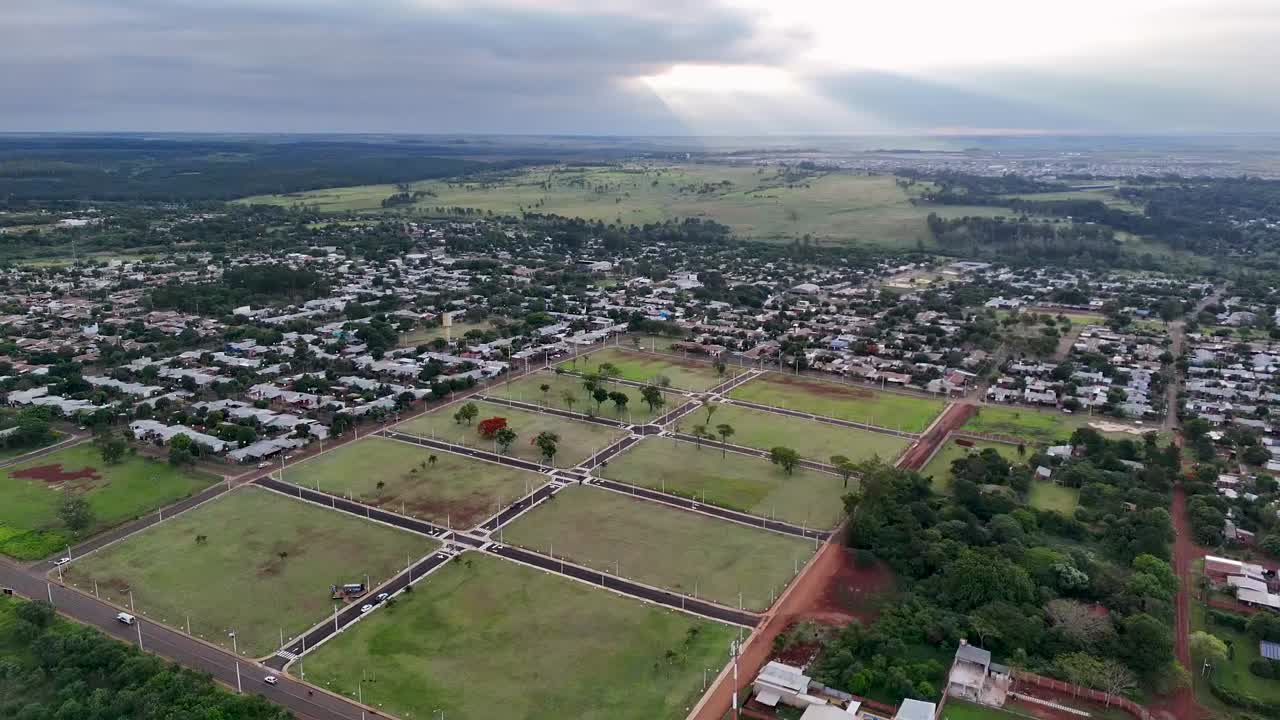 Drone pulling away over housing lots ready for construction in Posadas, Misiones, Argentina, revealing grid-like plots, surrounding neighborhoods, and open countryside under dramatic light
