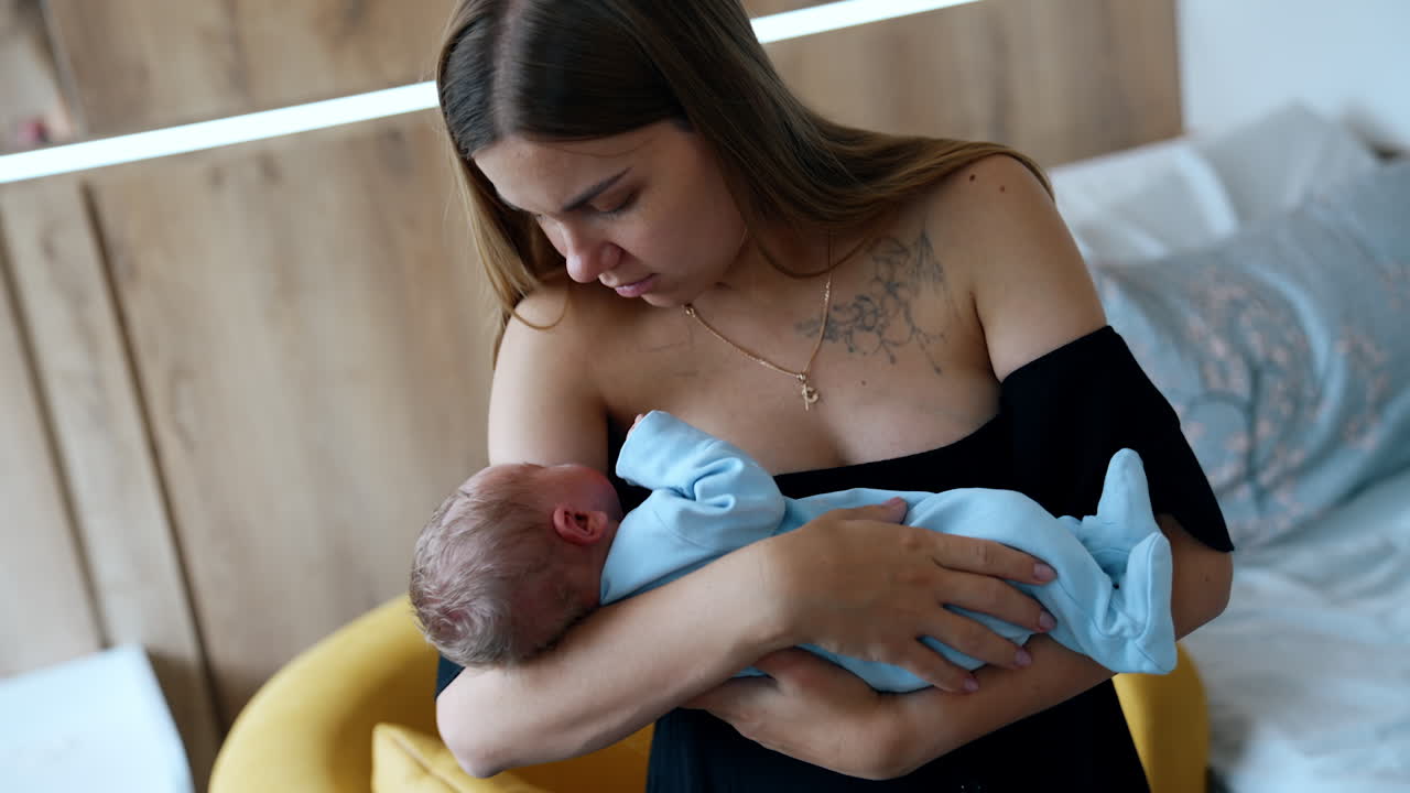 Loving Caucasian mom holding a newborn in hands. Mother is waving the child in blue romper.