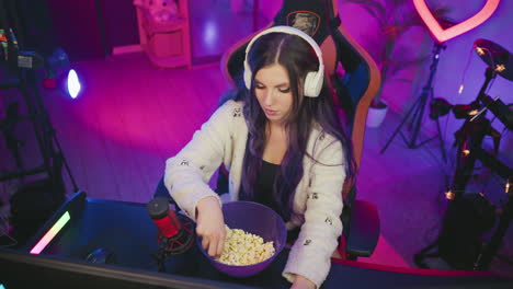 young female programmer wearing headphones types on computer in colorful studio, pauses to grab popcorn from bowl, then continues working in relaxed tech environment with glowing lights