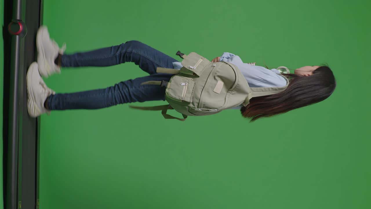 cuerpo lleno de vista lateral estudiante adolescente asiática con una mochila sosteniendo algunos libros y sonriendo mientras camina en la cinta de correr en el estudio de fondo de la pantalla verde