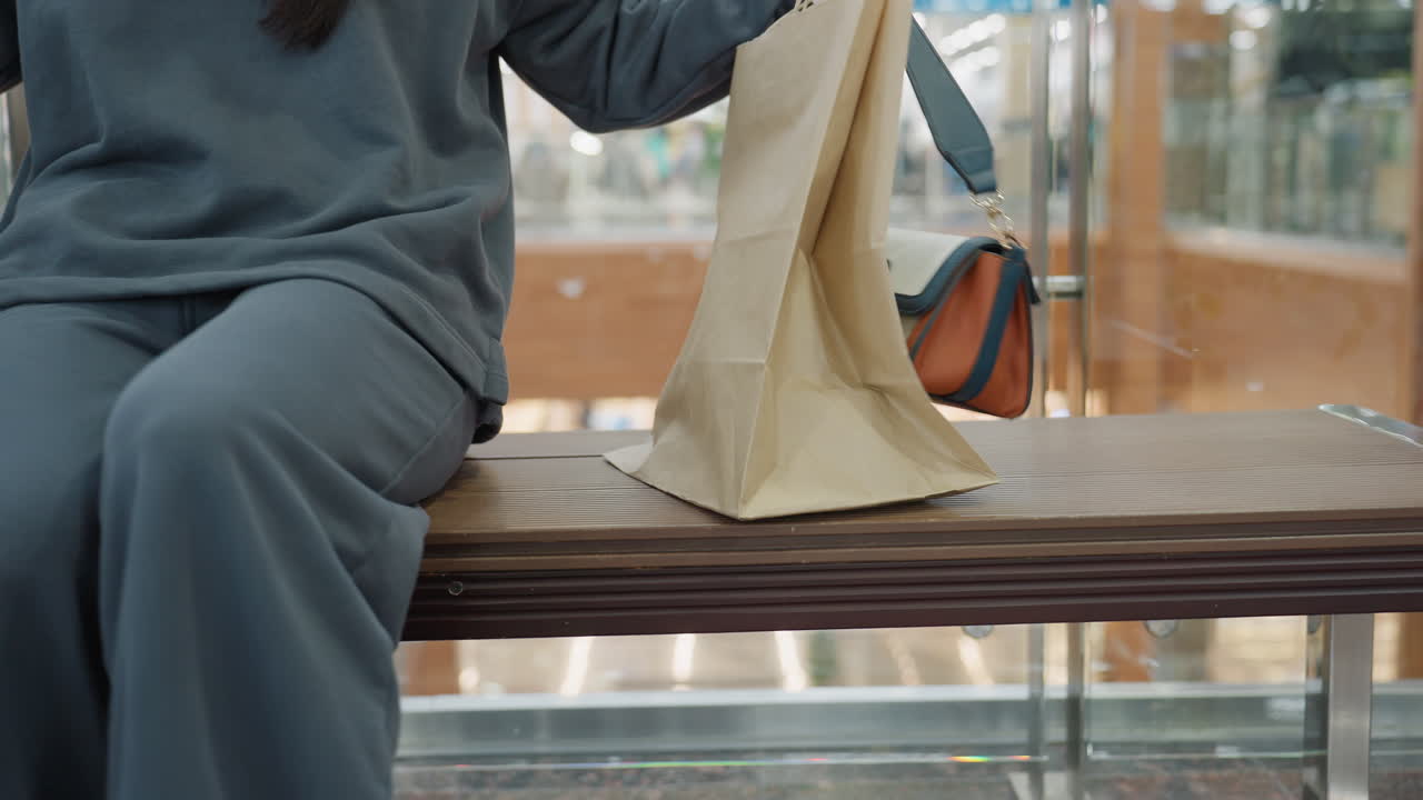 Partial view of shopper standing next to wooden bench in indoor mall while holding beige paper bag with orange handle in blur motion, glass barrier and retail background with colorful bokeh lights