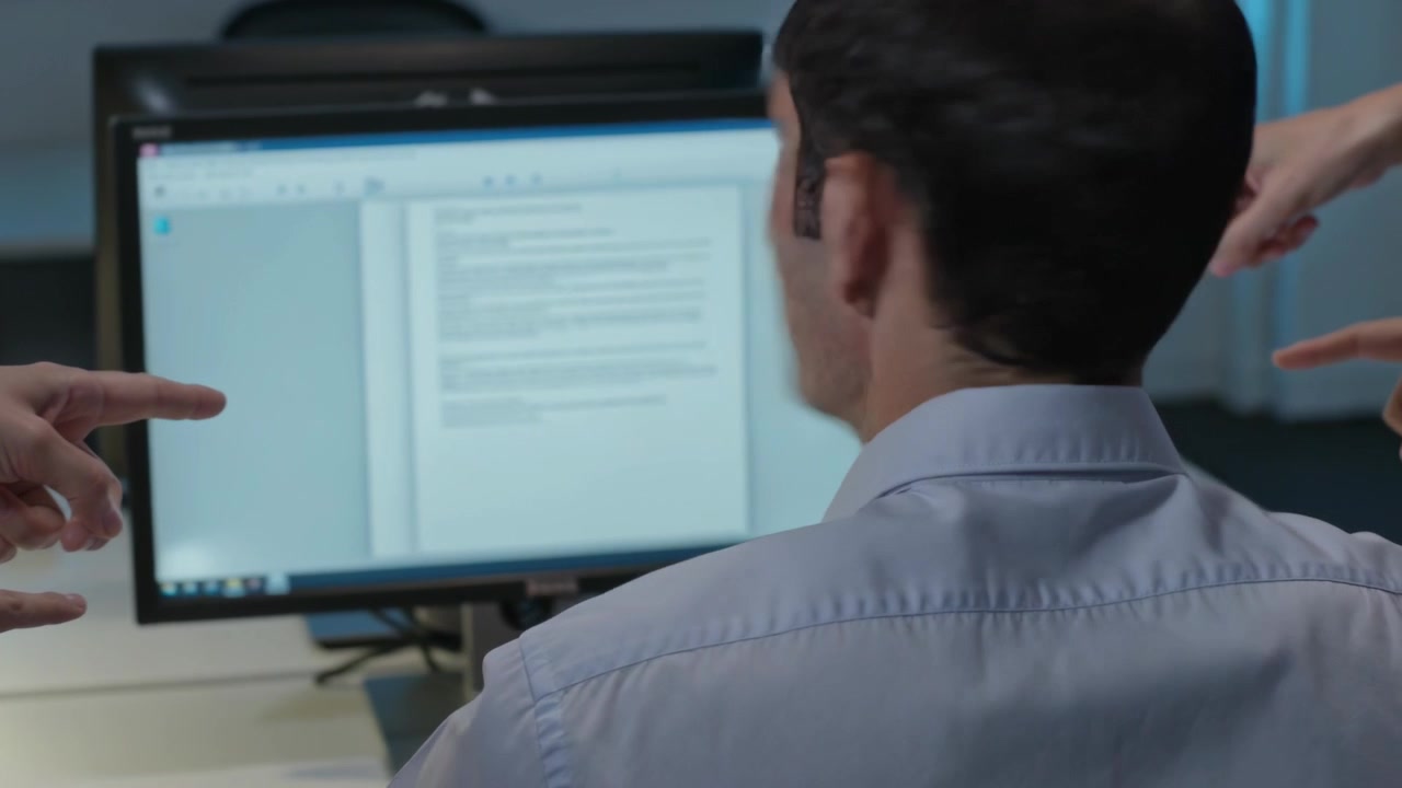 Mãos apontando fazem um homem de camisa azul clara se virar para o monitor no escritório, que exibe um documento.