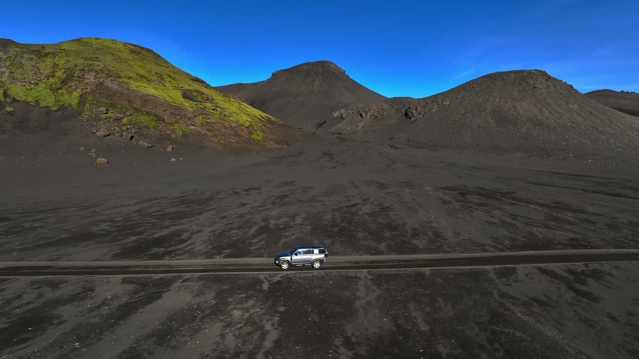 Ripresa laterale con drone di un SUV 4x4 che percorre una strada sterrata solitaria sotto colline vulcaniche nere e un cielo azzurro brillante. Avventura fuoristrada nelle remote alture con ampi spazi aperti